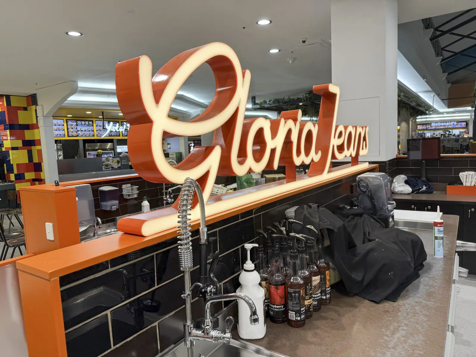 Illuminated Gloria Jean's script signage above the kiosk back-of-house bench, certified and signed off before opening